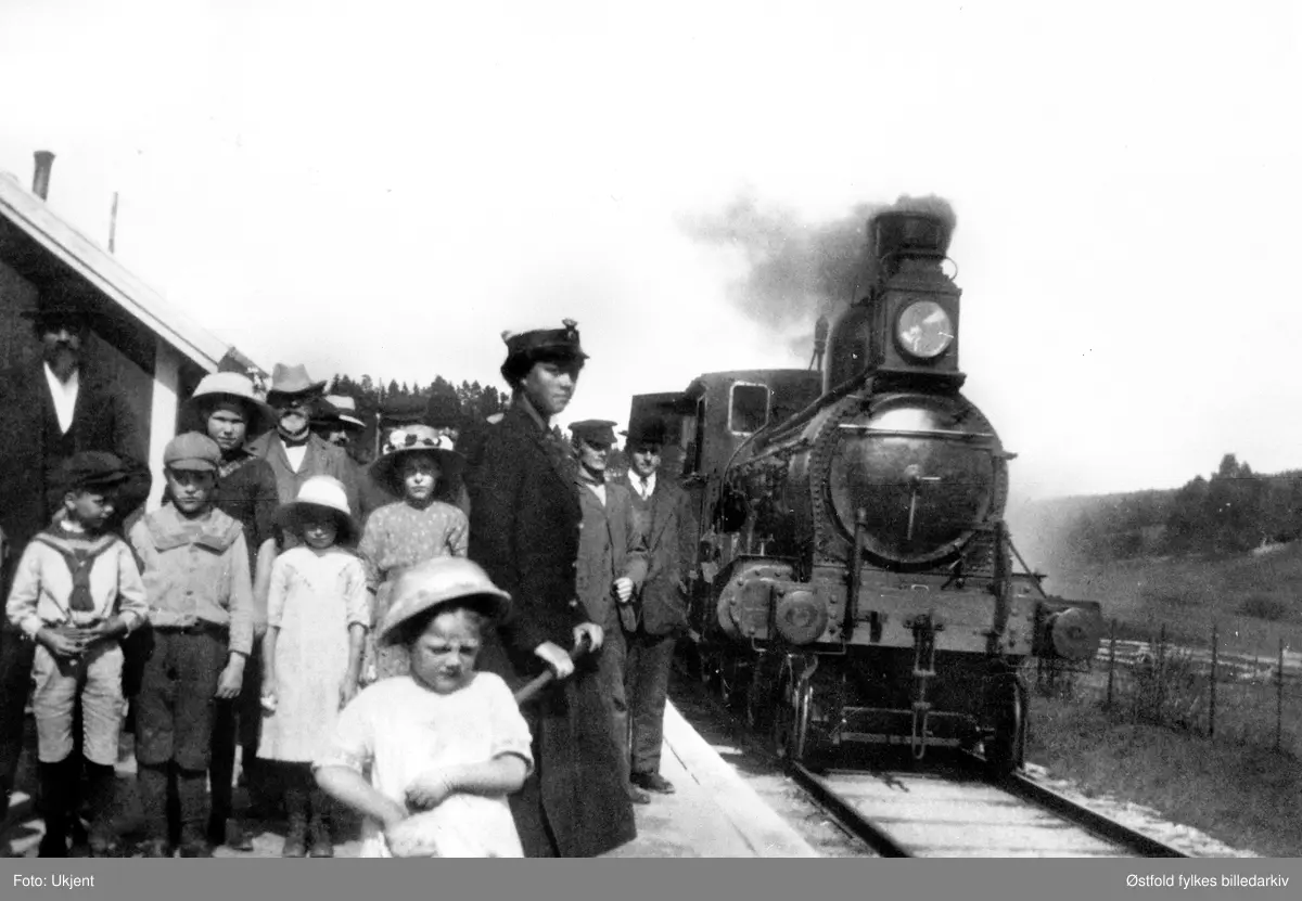 Jenny Elise Hansen (f. 18 nov. 1894) på Knapstad stoppested i Hobøl, østre linje, ca. 1913. Jenny var datter av gårdbruker og banevokter Johan Ludvig Furulund  (1871) og kona Nelly Emilie Furulund (f. Johansen), som dreiv gården Furulund i Hobøl . Jenny var muligens sesongansatt.
Stoppestedet Knapstad åpnet 15.11.1912, og stasjonsbygningen ble bygd 1915. Bygningen til venstre er antakelig  innkjøpslaget (bønder spleisa på å sette opp bygningen).
Per 1.10.1913 var Johan Ludvig Furulund banevokter og stasjonert på Tomter. Per 1918 var han blitt baneformann og stasjonert på Kråkstad.