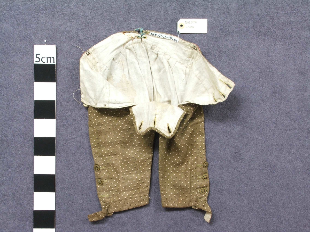 Jakke og bukse i bomullsstoff. Til gut. Høyrer saman som ei drakt.  Fôra med ufarga lin. Gul tråd i saum. Knappar i gull og svart. Buksa har klaff framme med tre knappar. Tre knappar nedst på kvart bein. Heilt nedst klaff med knapphol, her er knappane vekke. To knappar i lininga.  Knyting bak. Ein lapp i kvar side av lining. Jakka har silkeslag med påsydd pynt. Fire knapphol, berre ein knapp att. Fire hektar i heimelaga kvalitet til å hekta saman jakka øvst. På kvar side på jakka er det sydd på ein knapp med stoff.