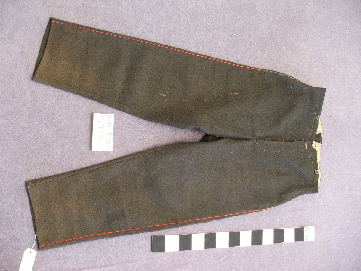 Uniformsbukse til mann. Svart med raud isydd stripe i sidesaumane. Fôra øvst ved lininga. Opning midt framme med fem knapphol. Ein knapp på kvar side av midten framme og bak. Kile isydd midt bak med eit lite hol på kvar side. Eit legg på kvar side av midtsaumen bak. Bak er påsydd to hemper i same stoff som buksa, hempene kan knappast saman. Metallknappar. Fire kilar isydd i skrittet.