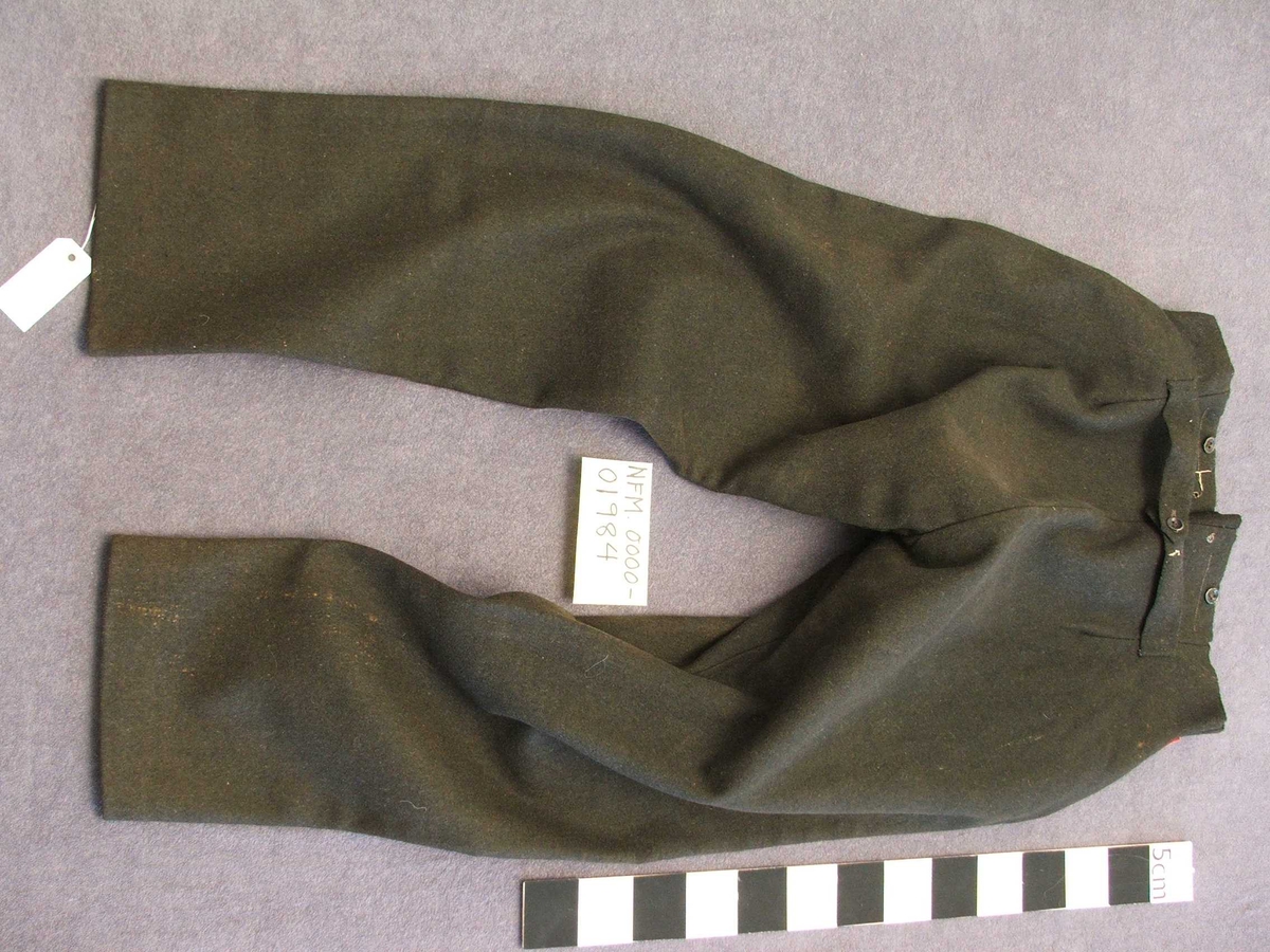 Uniformsbukse til mann. Svart med raud isydd stripe i sidesaumane. Fôra øvst ved lininga. Opning midt framme med fem knapphol. Ein knapp på kvar side av midten framme og bak. Kile isydd midt bak med eit lite hol på kvar side. Eit legg på kvar side av midtsaumen bak. Bak er påsydd to hemper i same stoff som buksa, hempene kan knappast saman. Metallknappar. Fire kilar isydd i skrittet.