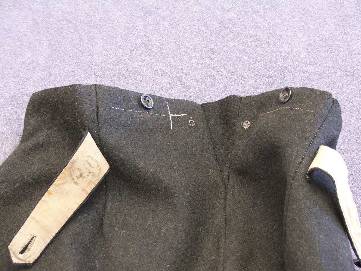 Uniformsbukse til mann. Svart med raud isydd stripe i sidesaumane. Fôra øvst ved lininga. Opning midt framme med fem knapphol. Ein knapp på kvar side av midten framme og bak. Kile isydd midt bak med eit lite hol på kvar side. Eit legg på kvar side av midtsaumen bak. Bak er påsydd to hemper i same stoff som buksa, hempene kan knappast saman. Metallknappar. Fire kilar isydd i skrittet.