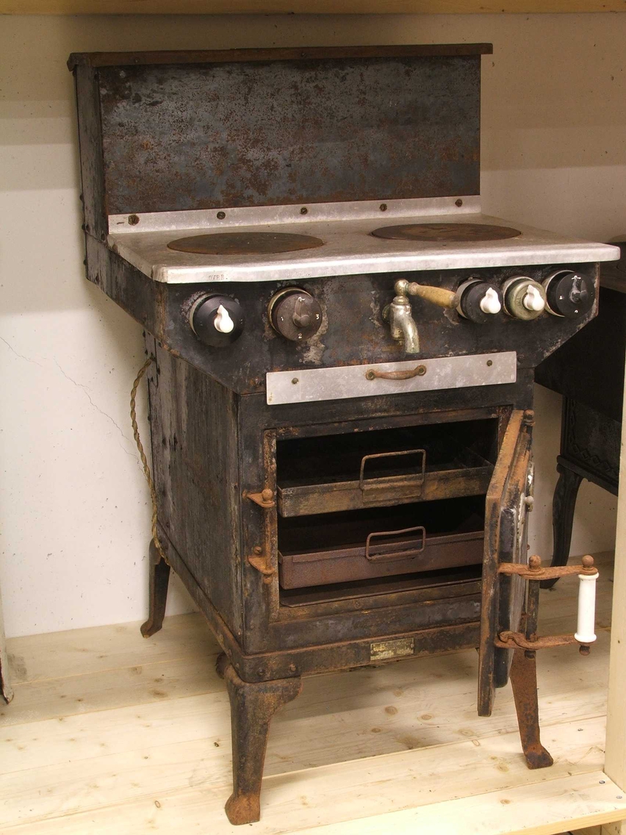 Flølo komfyren. Elektrisk komfyr. Med tank for varmt vatten (oppå bak), og kran for tapping av det varme vatnet framme. To kokeplater oppå. Fem runde brytarar framme i front. Ei uttrekkbar plate under. Omnsdør til å opne inn til steikjeomnen under der att. To plater med handtak inne i steikjeomnen. Komfyren står på fire bein (av jarn).