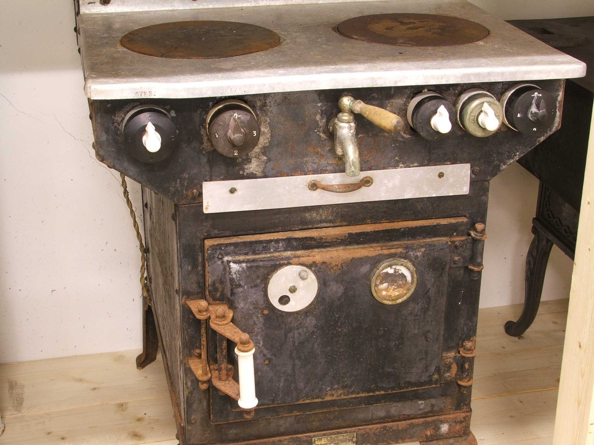 Flølo komfyren. Elektrisk komfyr. Med tank for varmt vatten (oppå bak), og kran for tapping av det varme vatnet framme. To kokeplater oppå. Fem runde brytarar framme i front. Ei uttrekkbar plate under. Omnsdør til å opne inn til steikjeomnen under der att. To plater med handtak inne i steikjeomnen. Komfyren står på fire bein (av jarn).