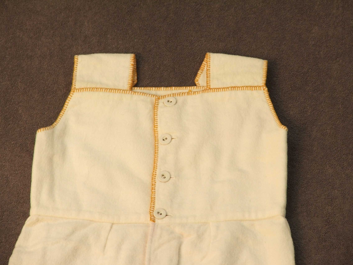 Den øvre delen er kanta med gule sting. Lerretsknappar; ein på kvart berestykke og for loddrett lukking 4 i øvre delen. Nedanfor knappane eitt stoffstykke som i kvar av dei to loddrette, innbretta sidene har 2 raude og ei blå stripe.