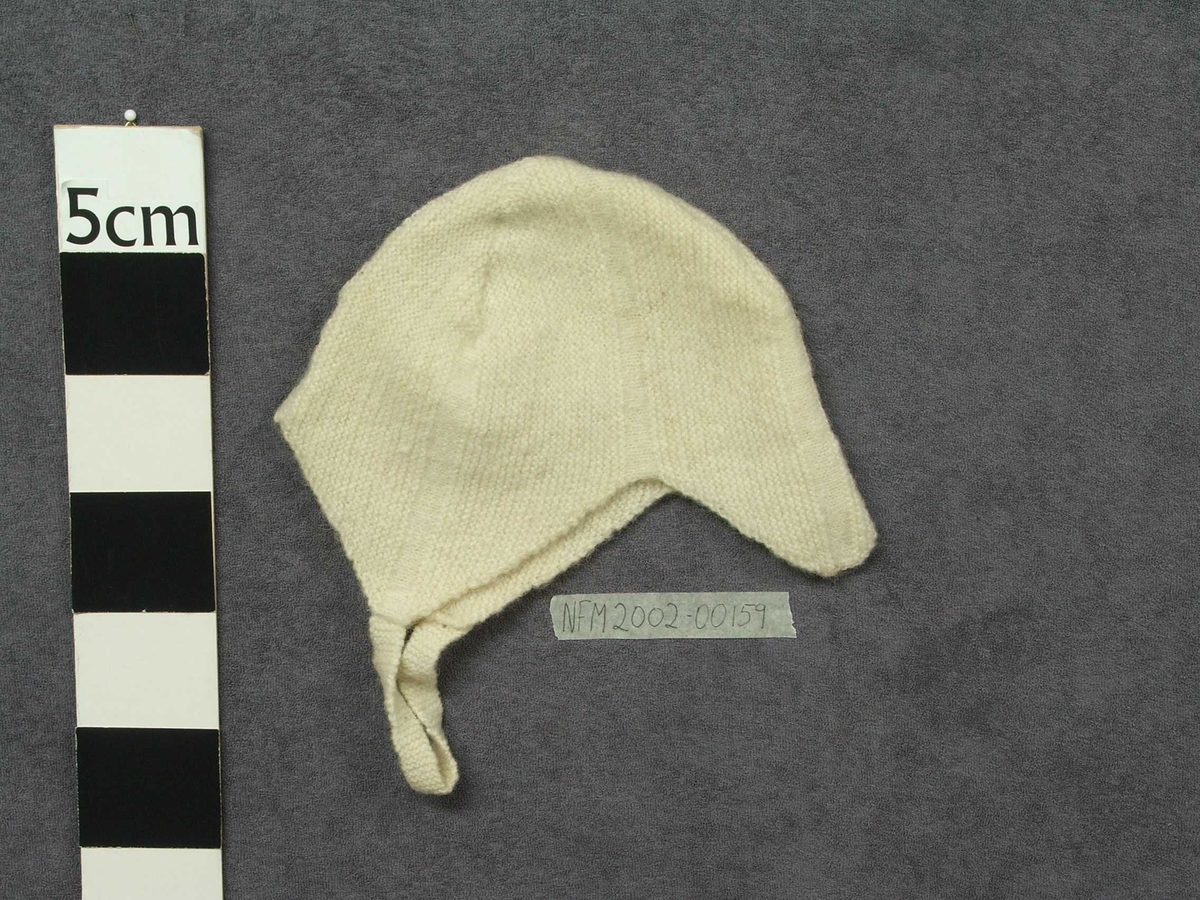 Strikka barnehue. Slags hjelm-fasong med ein spiss ned i panna. Strikka band til å ha under haka og knappe fast på andre sida.