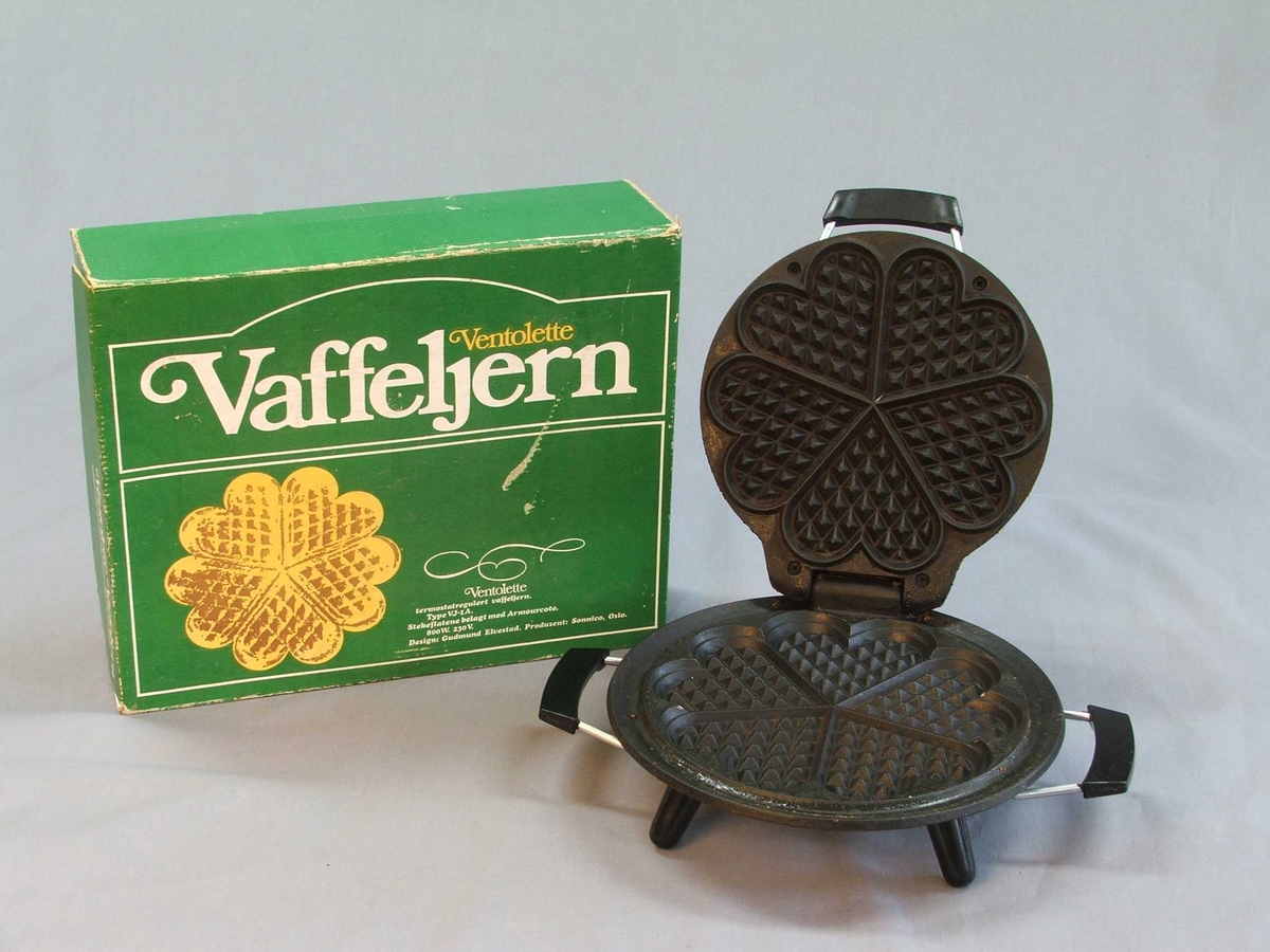 Eit vaffeljarn i original øsjke, isopor i øskja som jarnet står på. Det er ikkje ledning. Jarnet: Rund form. Loket opnast med handtak midt framme. Loket er hengsla fast til underdelen. Loket kan stå oppe av seg sjølv. Utsida av jarnet: oppå og under er det blankt metall, svart handtak på lok og to på kvar side av understellet. Inni er jarne svart, støypejarn? Vaffelhjarte-formar inni jarnet både på lok og botn = fem små hjarte som dannar ein sirkel. Fire svarte bein på undersida som jarnet står på.
Øskja er grøn med ein gul vaffel på oppsida og eit kvitt hjarte på sida. Øskja kan du opne framme. Den er kvit inni.