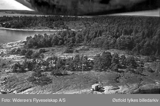 Skråfoto av hytter, m/flaggstang, sjøbod, brygge og båt. Fjell og skogsområder.
Bildet tatt nordover. Hytte beliggende vest for Brevik mot Bevø.