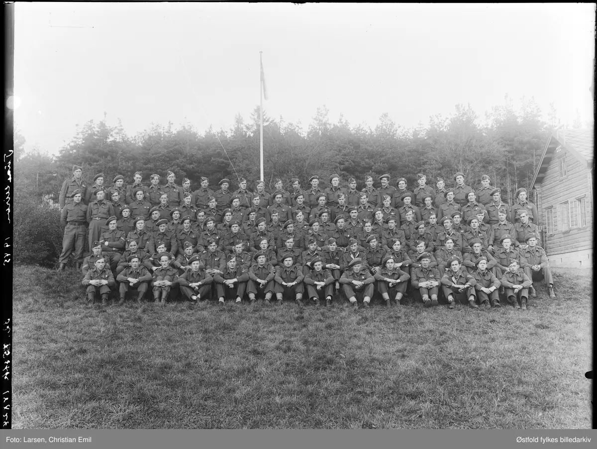 Tuneleiren i Tune 1945. Stort gruppebilde av soldater i uniform. Tuneleiren var tyskernes brakkeleir som lå på grunnen til Tune prestegård i Tune.
Man kan skimte «offiserboligen» i skogen helt til høyre på bildet, Tuneveien 46.