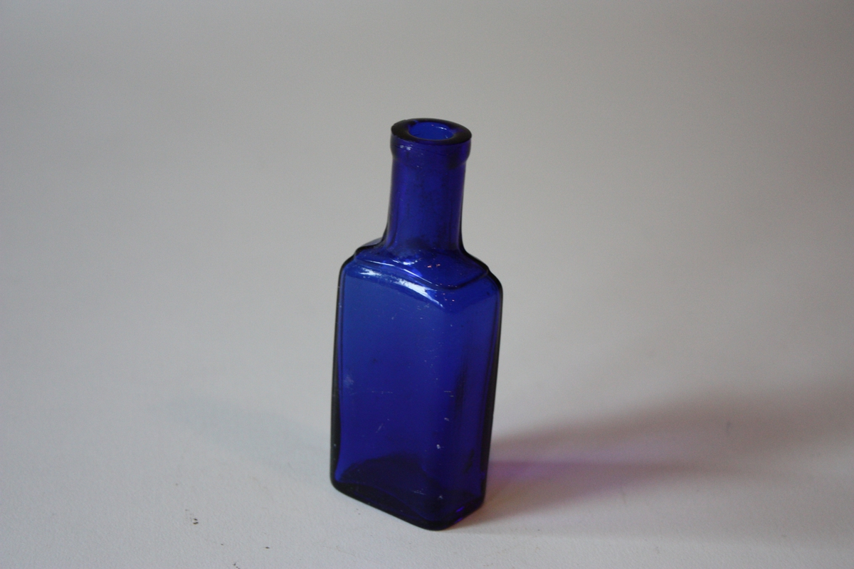 Flaske med rektangulært tverrsnitt, runda hjørne. Kraftig munning, har truleg hatt vanleg toppkork.