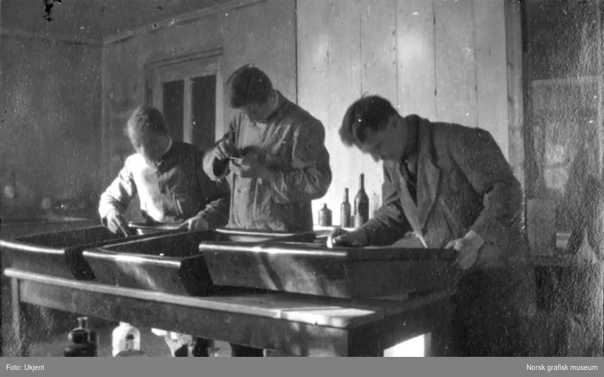 Tre menn står ved arbeidsbord med etsekar, interiør Dreyers Reproduktionsanstalt.

Albumtekst:
"Ætsning (amerikæn fæsjen)" - dvs. "på amerikansk måte".