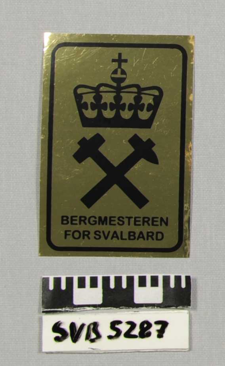 Firkantet klistremerke med trykt tekst og grafikk i gul og sort. Det er en hammer, en spisshakke og en kongekrone avbildet med tekst under.