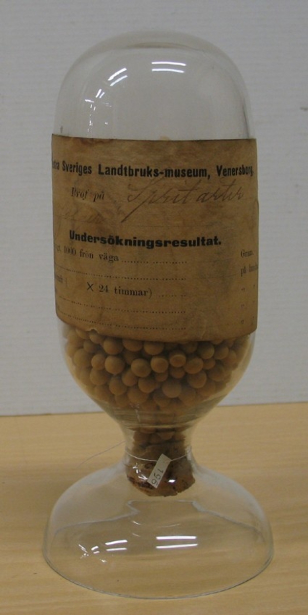 Glasbehållare innehållande fröer av spritärtor (Pisum sativum var sativum).

Ingår i en frösamling under namn: ''Westra Sveriges Lantbruksmuseum''. Bildat 1880 efter lantbruksmötet i Borås. Samlingen, vilken består av glasbehållare innehållande frön från svenska kulturväxter och träd, uppställdes först på länsresidenset i Vänersborg.

Samlingen kom att bli en av Vänersborgs museums 4 grundsamlingar och uppställdes i museets övre våning vid museets öppnande 1891.

Troligen är glasbehållaren tillverkad vid Reijmyre glasbruk, ligger i Rejmyre, norr om Norrköping.