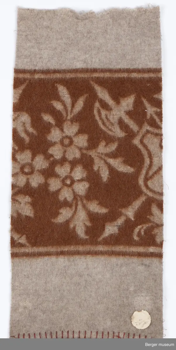 1 prøve. Teppet er ensfarget med en bred bord. I borden er det blomster og et våpenskjold med pil og øks.  Art Nouveau.