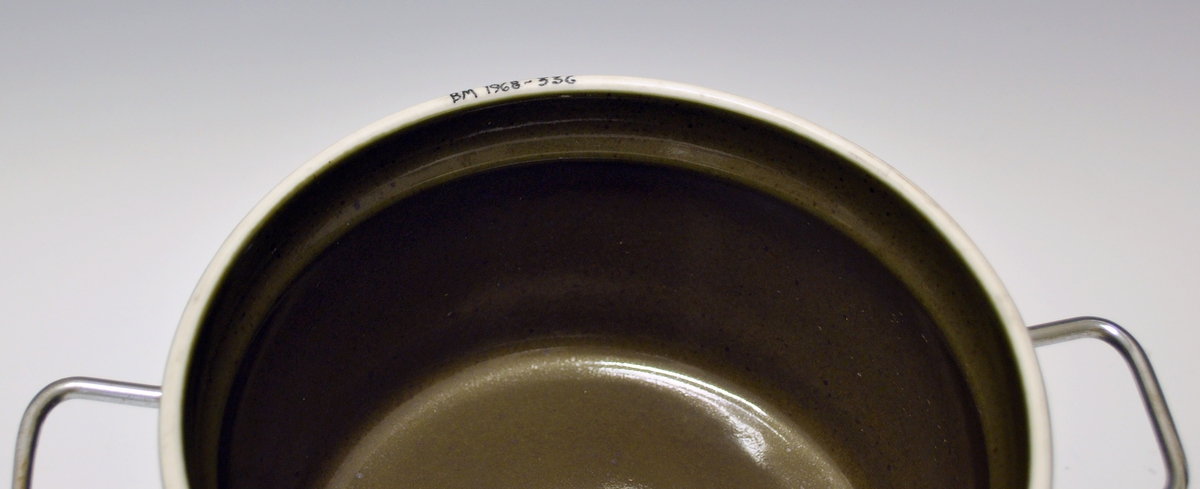 Gryte med lokk av porselen, ildfast gods. Mørkegrønn glasur. Metallhanker.
Modell 2376 Askeladden av Eystein Sandnes, i produksjon fra 1964.
