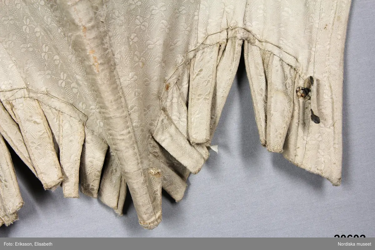 Huvudliggaren:
"Snörlif. Af hvitt siden, från 1770-talet."

Liv till hovdräkt s.k. "robe á la cour" av ursprungligen vit sidendroguet. Tyget förmodligen svensk tillverkning (Myrman?) se 1700-tals textil, A. Berchs samling, sid 171. Yttertyget är skuret i nio delar med mittstycke fram som löper ned i snibb samt fyra delar å varje sida med snörning mitt bak. Grundlivet tillverkat av oblekt linnelärft är helt styvat med spröt av valben trädda i sydda vertikala kanaler samt med en skena av järn som löper runt urringningen och planschett mitt fram. Grundlivet troligen klistrat med papper för att hålla en viss form innan yttertyget monterats på. Ärmholkar bestående av å varje sida tio hopsydda delar som lagts i veck. Tillskärningen följer helt den gängse för robe de cour. Se Waugh, Nora, The Cut of Womens Clothes, s. Längs sidorna i midjehöjd en tunn snodd avsedd att haka fast släpet i. Livet ursprungligen buret med kjol och släp i samma tyg.
/Ingrid Roos 2007-03-06