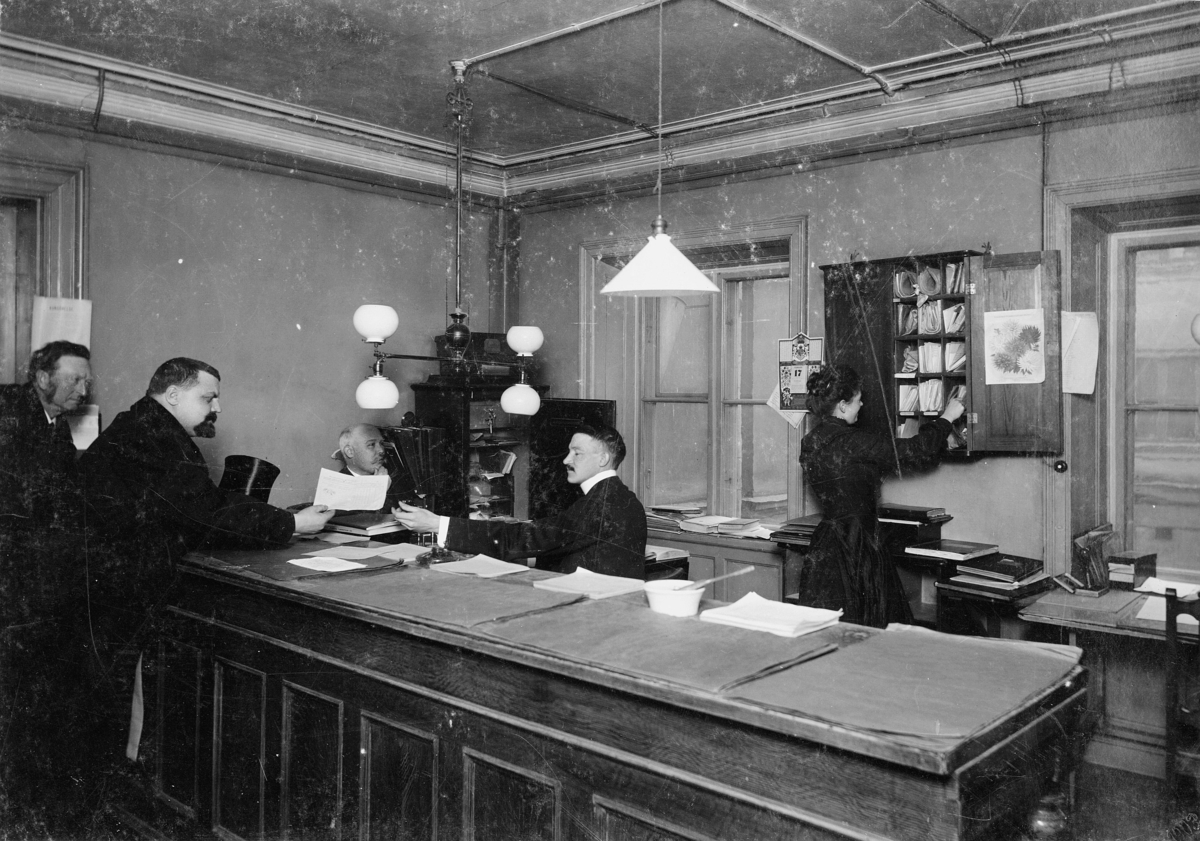 Posthuset Rödbodtorget, Stockholm 1903.
Postkontoret Stockholm 1.  Tidningsexpeditionens kassa exp.
Personalen fr.v.: P? Lundholm,  J.L. Hallmén,  E. Söderholm.
