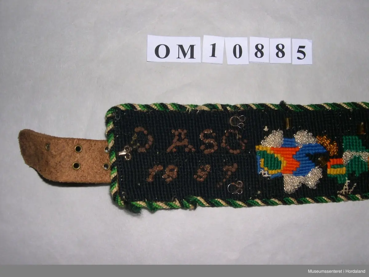Brodert mannsbelte. Blomstermotiv med perledekor. Svart hovudfarge, blomster i grønt, gult, blått og oransje. Blanke og gullfarga småperler, nokre store, korte rørperler i gullfarge. I eine enden er det initialar og årstal, OASG 1887, sydde med perler som har rusta. Tvinna, fleirfarga snor til kanting. Raudt lêr til bakstoff. Beltet har beltespenne på baksida litt inn på beltet, og skinnreim med hol i andre enden. I tillegg er det fleire maljer og hekter.