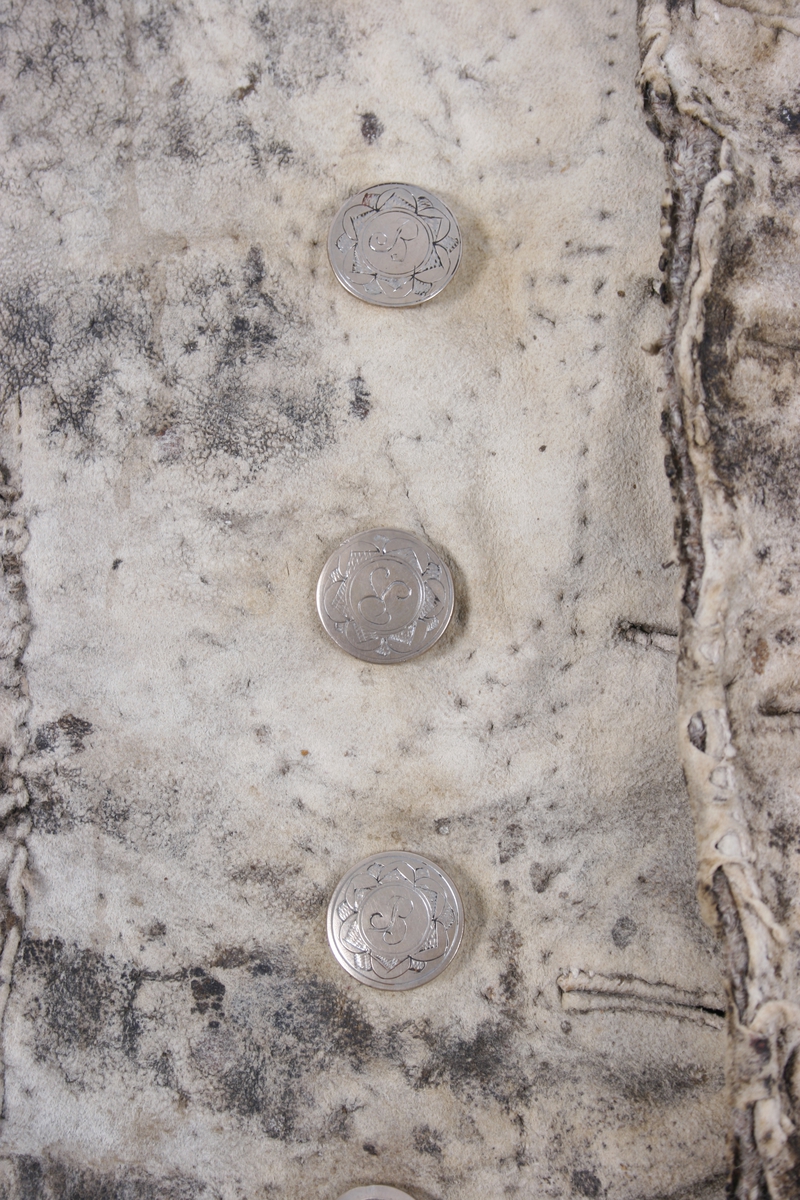 Dobbeltknept skinnvest med 9 knapper og passepoilerte knapphull på hvert framstykke. Knappene er festet med en skinnreim på innsiden . Den er sydd fast til foret. Øverste knapphull på høyre framstykke er sydd med knapphullsting. Knappene er siselerte med bokstaven B i midten med en bord rundt.  Den nederste knappen på begge framstykkene er mindre enn de andre og har ikke samme mønster som de andre. Vesten har 1 brystlomme og 2 lommer i livhøyde. 1 innerlomme i skinn er sydd fast til foret. Foret er sydd sammen av mange små stoffbiter i 2 farger. Foret og skinnet er stukket sammen. Bakovertrekte sidesømmer. Påsydd skinnkant rundt halsrigning og langs begge sider MF. Framstykkene går ned i en spiss MF.