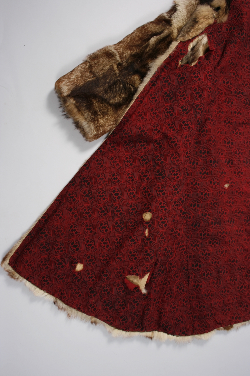 Ulveskinnspels, Lang og noe utskrådd. 
Sydd sammen av mange smale pelsstykker, to i høyden. En liten splitt midt bak. Ermer sydd sammen av mindre pelsstykker. Ermene har en liten oppbrettet fald av pelsen i nedkant, på resten av plagget er pels og fór sydd sammen kant i kant, ute belegg eller oppbrettet fald. Sjalskrage med underkrage av sort kunstpels. Underkragen har søm midt bak. 
Fóret med rødt klede med sort trykk, også i ermene.
Hele plagget er håndsydd med grov tråd, selve pelsen med ubleket lintråd. Fóret er sydd sammen, og sydd til pelsen, med ubleket eller sort bomullstråd. Fóret har sidesømmer, men ingen søm i ryggen. Ryggstykket er kvadratisk med trådrette sider. Forstykkene er trådrette midt foran og skrådd i siden. I sidene er det satt inn en liten kile nederst. 
I nedkant av kragen i venstre side er det en stor sølvhekte, gravert med blomstermotiv. Ingen tegn til annen lukning.  
Stor lærhempe i nakken.