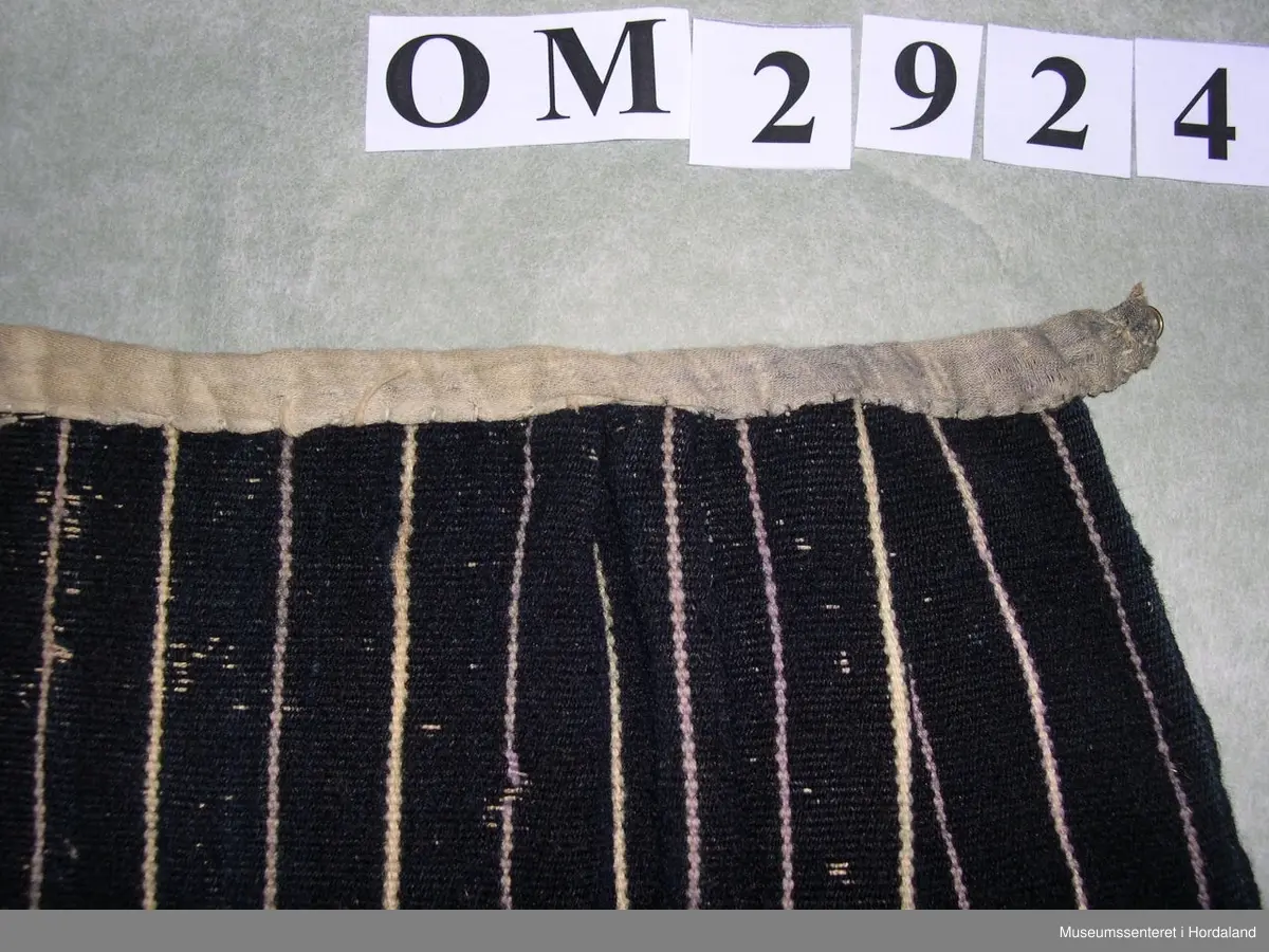 Mørkeblått forkle med kvite og lyselilla, smale og jamne striper. Linning av silkeband. To ringar med hyssing. Framme er det glatt parti og to folder på kvar side.