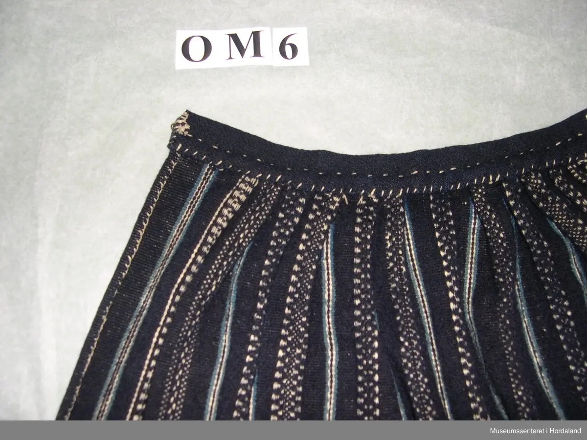 Mørkeblått, vove, stripete forkle i ull. Stripene er kvite og lyseblå. Linning med ein ring i kvar ende for feste av forkleband. Smalt, glatt stykke framme med seks folder på kvar side.