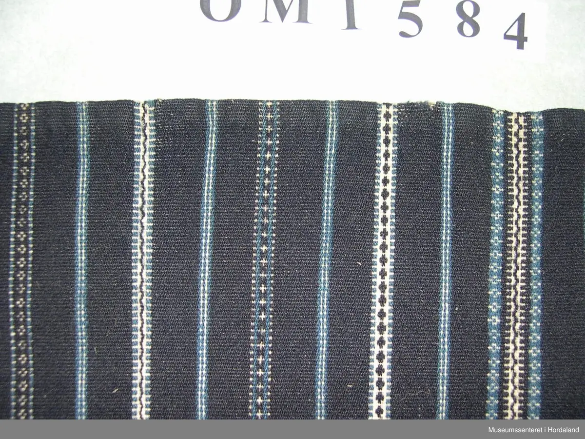 Mørkeblått, vove ullforkle med striper i kvitt og mellomblått. Linning. Glatt stykke midt framme, to folder på kvar side. Ei hekte til attlat.