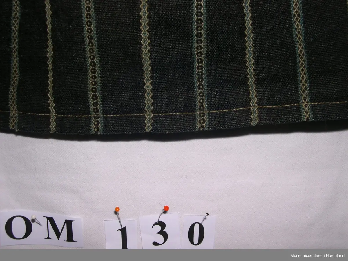 Mørkeblått, stripete vove forkle i ull. Striper i kvitt og blått mønster. Linning. Glatt stykke midt framme, tre folder på kvar side. Brunt fløyelsband nede. Skoning på baksiden med mønstra bomullsstoff og saum som syner på framsida. Forkle er smalt oppe. Holete.