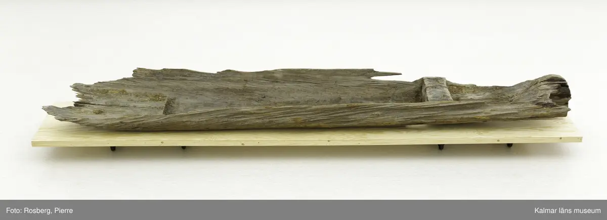 KLM 9303. Båt, stockbåt. Av urholkad trädstam. Huggen i ett stycke. Den har subrektangulärt tvärsnitt, dvs. rektangulär form med rundade kanter. Båten är formad med ett mittre djupare rum med högre för- och akterpartier. Mittpartiet är avgränsat med vertikala tvärväggar, skottar, som utgörs av den bibehållna stocken. Den bakre tvärväggen är högre och har troligen använts som toft. I aktern en köl som sträcker sig fram till ungefär mitten av båten. Framför tväravsatsen i aktern, toften, finns babord om kölen ett rektangulärt hål 15 x 6 cm i botten. På båten en beläggning, troligen sediment från sjön den hittades i.