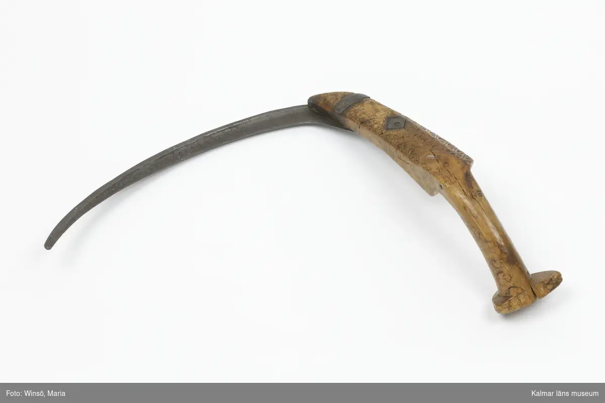 KLM 7163. Skära, vinkelskära. Av trä och metall. Vinkelskära med bågböjt blad av järn, fastnitat i skaftet. Skaft av masurbjörk, ornerat med plattsnidat rutmönster. Ornerat vågmönster på bladets ena sida samt inpunsat: ANO 1788 SIS. Rund knopp, en bit saknas. På knoppen skuret sicksackmönster samt 1788 SIS. Användes för att skära av säden.