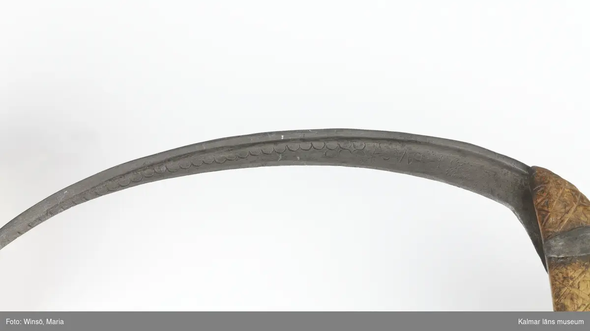 KLM 7163. Skära, vinkelskära. Av trä och metall. Vinkelskära med bågböjt blad av järn, fastnitat i skaftet. Skaft av masurbjörk, ornerat med plattsnidat rutmönster. Ornerat vågmönster på bladets ena sida samt inpunsat: ANO 1788 SIS. Rund knopp, en bit saknas. På knoppen skuret sicksackmönster samt 1788 SIS. Användes för att skära av säden.