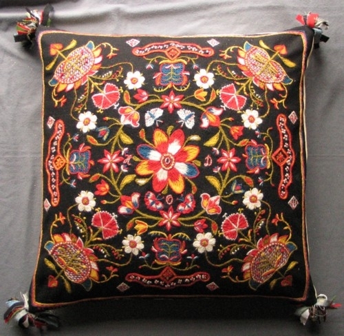 Broderad kudde i Skånskt yllebroderi. Svart botten, sämskskinn på bakstycket och duskor i hörnen. Liten blomkrans med blomma och fyra fjärilar i mitten. I hörnen solrosor med utskjutande bukett. På sidorna låsornament.   Kudden är sydd mestadels med klyvsöm. Orginalet kommer från Gälltofta utanför Rinkaby, Kristianstad, och finns nu på Regionmuséet i Kristianstad.