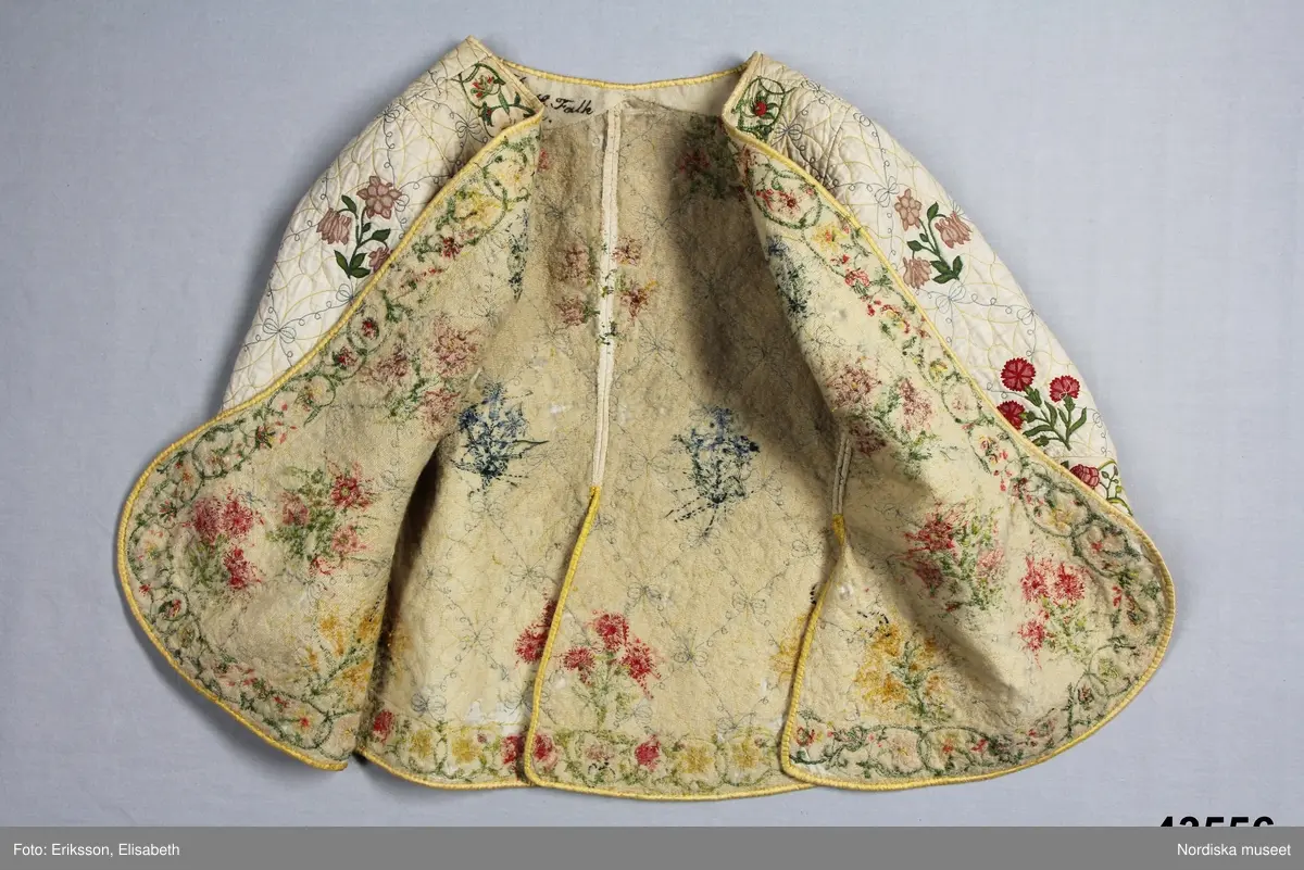 Katalogkort IV:
"Tröja / kofta, för dam, 1600-talets slut.
Längd 54 cm
Av linnelärt med foder av glest tuskaftat ylletyg. Försedd med stickningar i gult och blått samt broderier i kedje-söm med yllegarn. 2 ryggstycken, 2 framstycken. Halvlånga ärmar med 2 sömmar, varav den ena öppen och kantad med gult siden samt försedd med gult silke tränsade snörhål, med band av dubbelvikt sammansytt band. Sömarna uppslitsade nedifrån upptill midjan och kantade med gult siden."