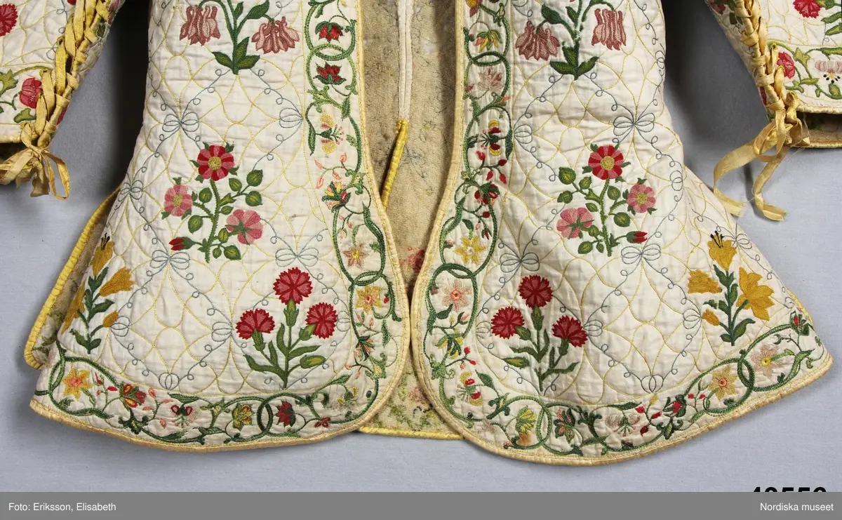 Katalogkort IV:
"Tröja / kofta, för dam, 1600-talets slut.
Längd 54 cm
Av linnelärt med foder av glest tuskaftat ylletyg. Försedd med stickningar i gult och blått samt broderier i kedje-söm med yllegarn. 2 ryggstycken, 2 framstycken. Halvlånga ärmar med 2 sömmar, varav den ena öppen och kantad med gult siden samt försedd med gult silke tränsade snörhål, med band av dubbelvikt sammansytt band. Sömarna uppslitsade nedifrån upptill midjan och kantade med gult siden."