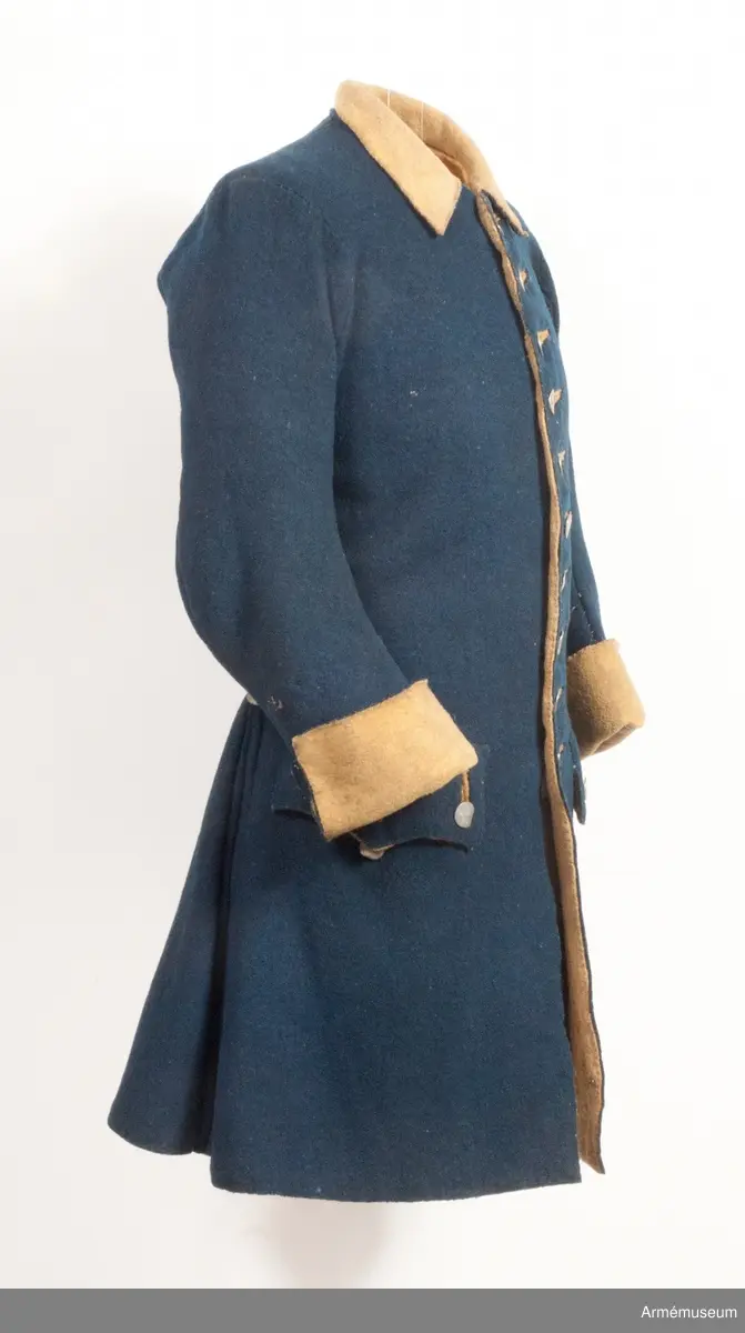 Rock av blått kläde med gul krage och ärmuppslag. Fodrad med gul boj i livet och med linne i ärmarna. Försedd med tennknappar.