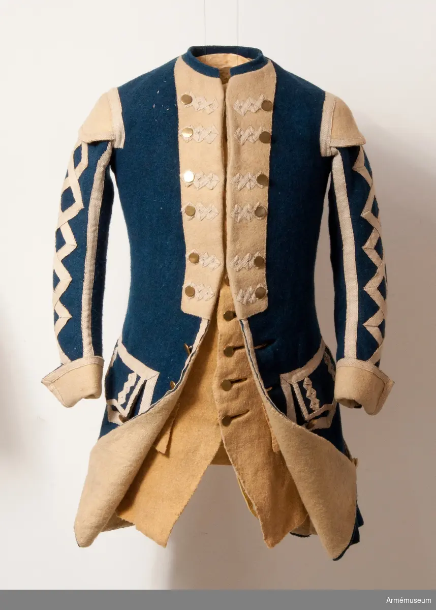 Rock med prydnader för spelet; mellanblå med gul rabatt (bröstrevärer), svalbon (axeldekoration) och ärmuppslag. Vit redgarnsdekor bruklig för spel. Rocken har sammanlagt 23 knappar. Knapparna i framkanten fastträdda med vitt band från avigsidan. En knapp i vardera skört (en ny). Två knappar på ficklocken (höger sida fattas en). Rocken är fodrad med gult ylletyg. Rocken ihopfästes med hyskar och hakar.