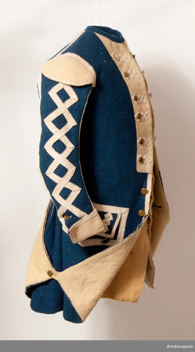 Rock med prydnader för spelet; mellanblå med gul rabatt (bröstrevärer), svalbon (axeldekoration) och ärmuppslag. Vit redgarnsdekor bruklig för spel. Rocken har sammanlagt 23 knappar. Knapparna i framkanten fastträdda med vitt band från avigsidan. En knapp i vardera skört (en ny). Två knappar på ficklocken (höger sida fattas en). Rocken är fodrad med gult ylletyg. Rocken ihopfästes med hyskar och hakar.