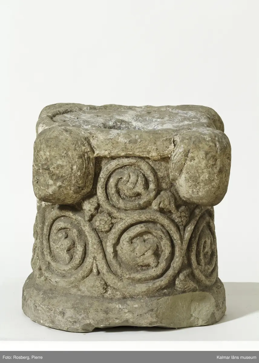 KLM 35314. Dopfunt, dopfuntsfot. Av kalksten. Fot till dopfunt med rund genom skärning. Spiralupprullade växtrankor i relief samt upptill fyra kraftigt utstående ansiktsmasker. Ett cylindrisk hål tvärs igenom stenen.