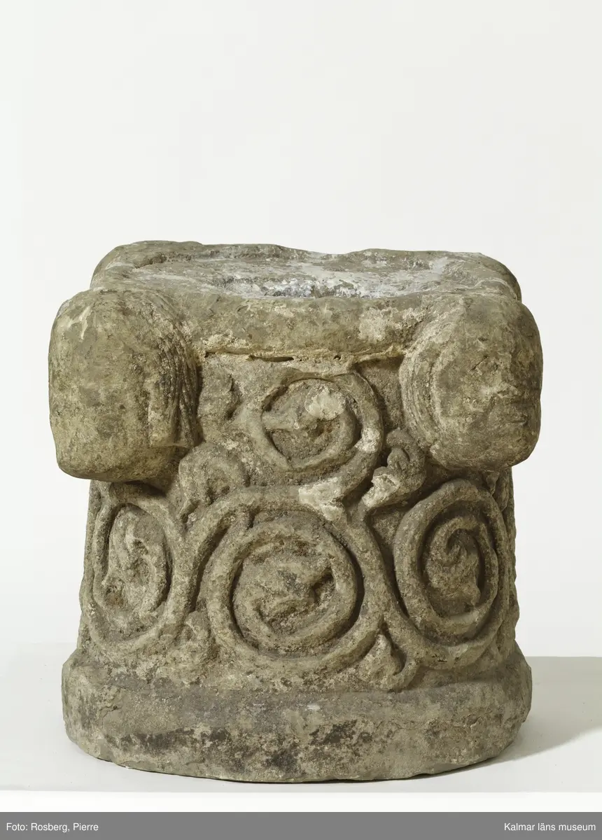 KLM 35314. Dopfunt, dopfuntsfot. Av kalksten. Fot till dopfunt med rund genom skärning. Spiralupprullade växtrankor i relief samt upptill fyra kraftigt utstående ansiktsmasker. Ett cylindrisk hål tvärs igenom stenen.