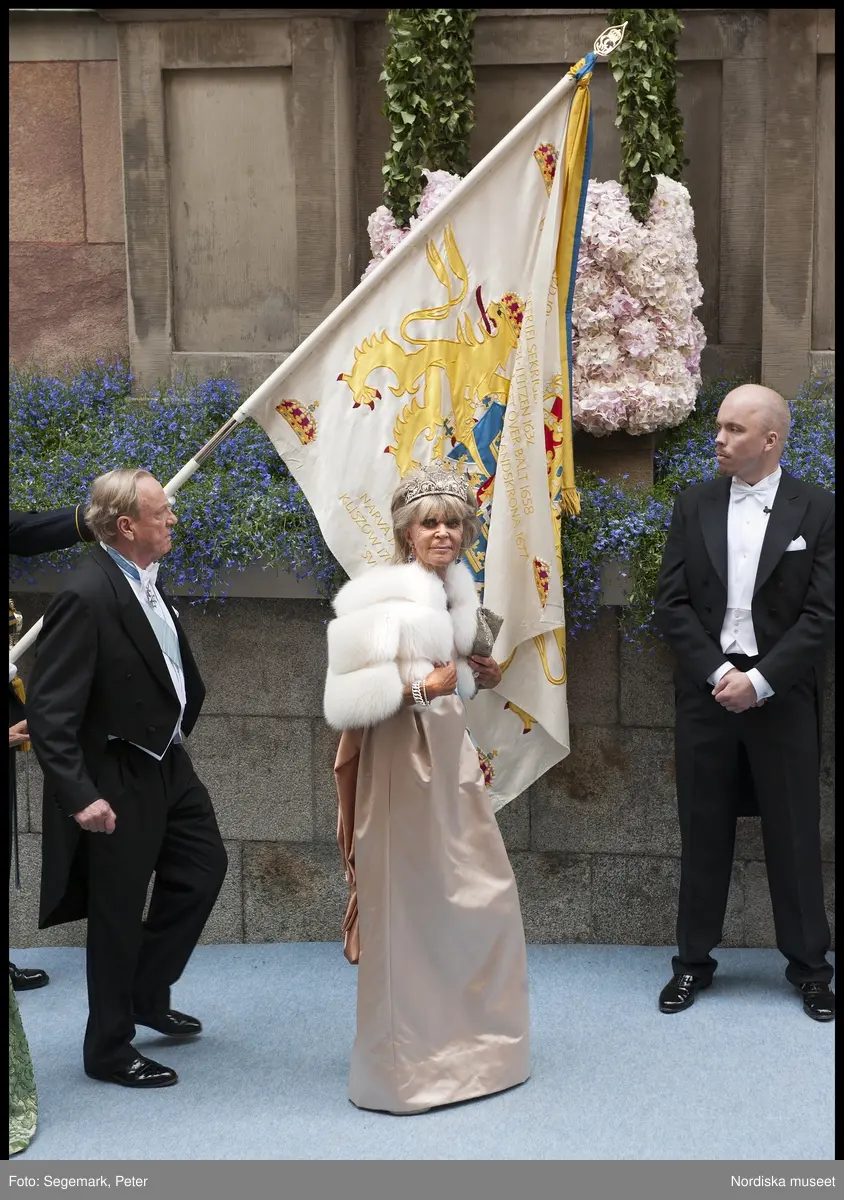 Kronprinsessbröllopet. Dokumentation av bröllopet mellan Kronprinsessan Victoria och herr Daniel Westling 19 juni 2010. Gästerna anländer till Storkyrkan. Prinsessan Birgitta i champagnefärgad sidenklänning med vit pälsjacka, drottning Sofias diadem, smycken, kuvertväska och ordenstecken. Make, Johan Georg av Sigmaringen i frack och ordenstecken.