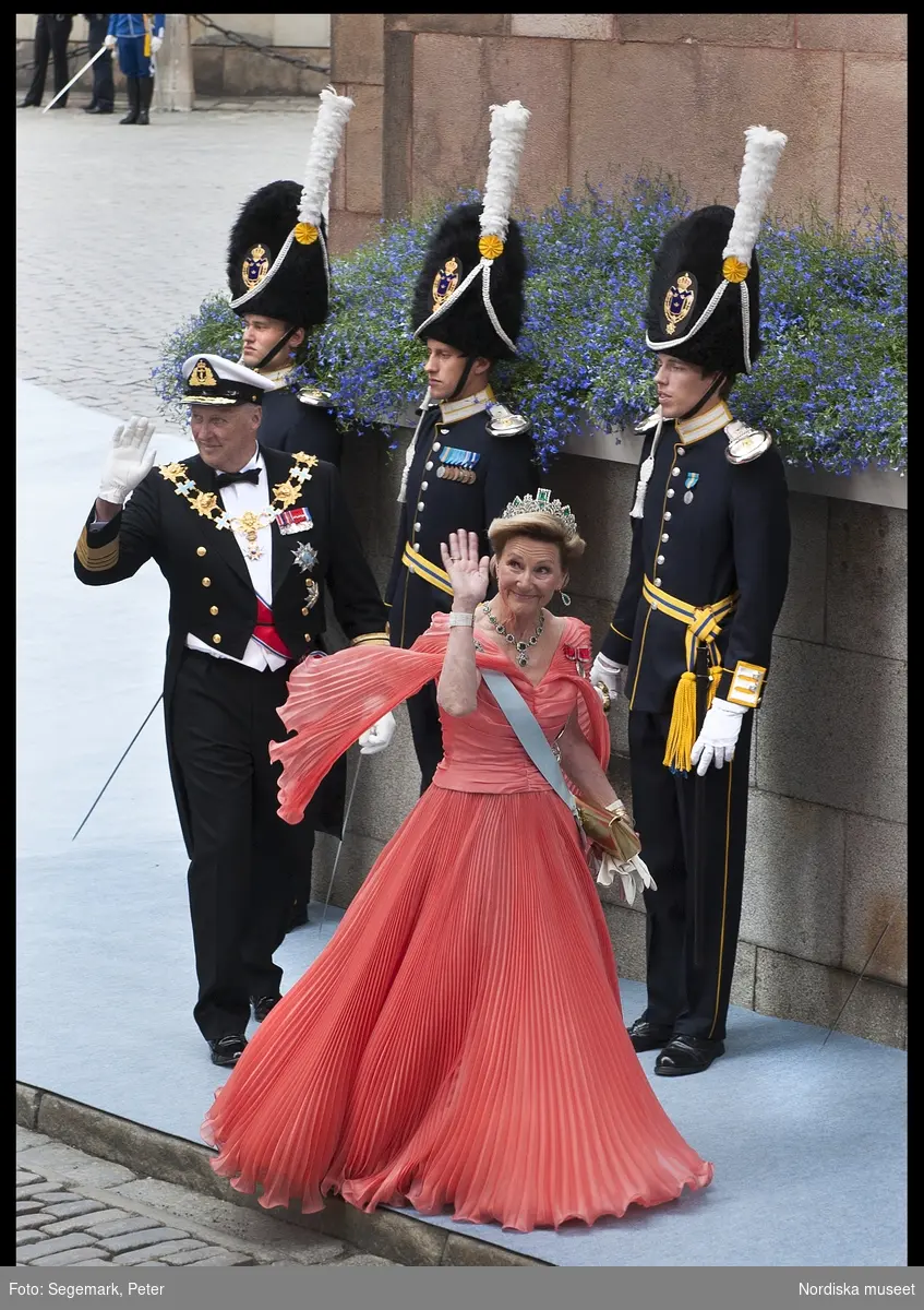 Kronprinsessbröllopet. Dokumentation av bröllopet mellan Kronprinsessan Victoria och herr Daniel Westling 19 juni 2010. Gästerna anländer till Storkyrkan. Kung Harald av Norge i stor mässdräkt och ordenstecken med makan drottning Sonja i aprikosfärgad, helplisserad, sidenklänning och ärmcape i samma nyans, diadem, smycken, guldfärgad kuvertväska och ordenstecken.