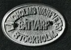 Varvskylt, mässing, gjuten.
Text: Långholmswarfvets Båtvarf Stockholm.
Längd: 7cm. Bredd: 4,7 cm.