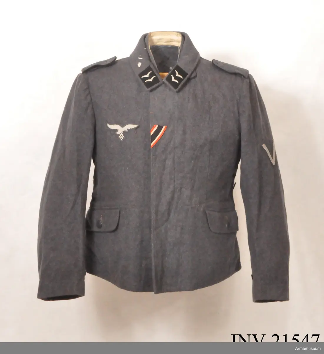 Av fältgrå kläde. Med knäppjulp. Axelkklaffar av samma grå kläde med svart passpoil. Foder av grått bomullstyg med två innerfickor. Knappar av grå metall. Krage, öppen, nedviken med klaffar av svart kläde på kragens ända.