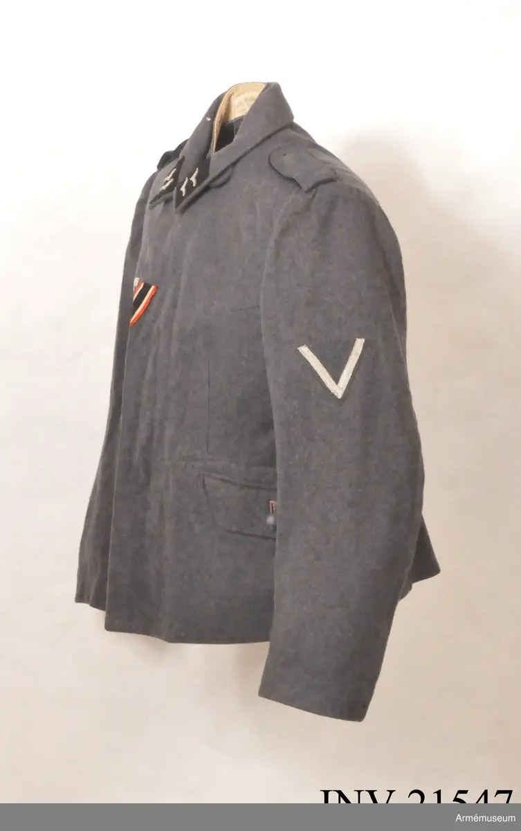 Av fältgrå kläde. Med knäppjulp. Axelkklaffar av samma grå kläde med svart passpoil. Foder av grått bomullstyg med två innerfickor. Knappar av grå metall. Krage, öppen, nedviken med klaffar av svart kläde på kragens ända.