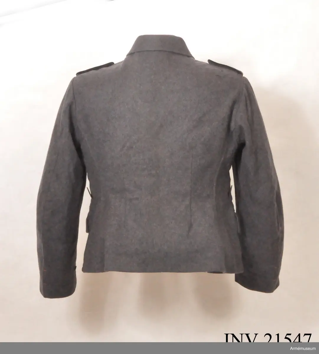 Av fältgrå kläde. Med knäppjulp. Axelkklaffar av samma grå kläde med svart passpoil. Foder av grått bomullstyg med två innerfickor. Knappar av grå metall. Krage, öppen, nedviken med klaffar av svart kläde på kragens ända.