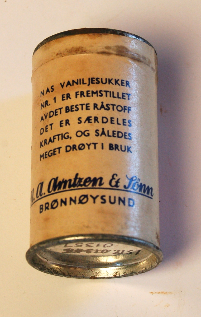 Sylinderformet blekkboks med påklistret papiretikett med varedeklarasjon: "NAS vaniljesukker Nr.1 er fremstillet av det beste råstoff. Det er særdeles kraftig, og således meget drøyt i bruk".