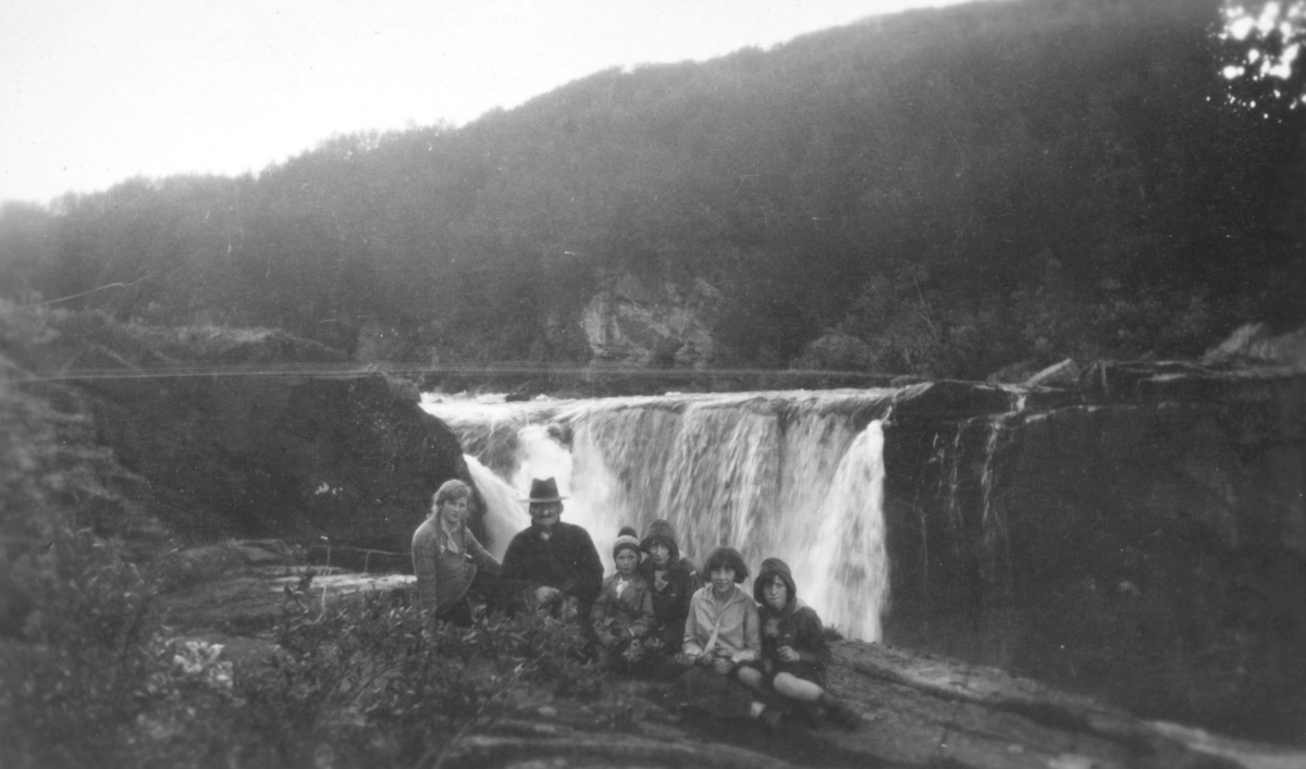 Bildet er tatt ovenfor Bergeby i juli 1930. En gruppe mennesker poserer forran en foss. I bakgrunnen er det et fjell. Fra venstre sitter Ingebjørg Fosslund, Henrik Henriksen, Brit Hornburg, Dagny Nordahl, og Johanna Dahl