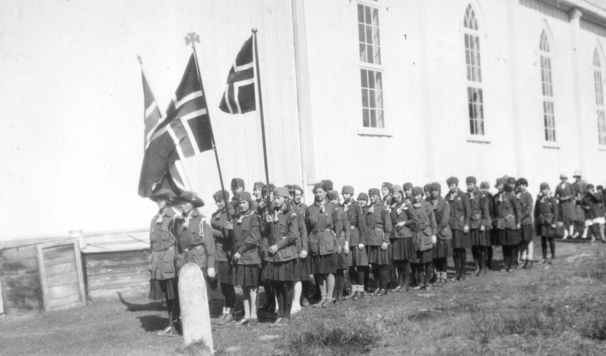 Speiderjenter på Kretsleir i Alta, 1928. Her står de i tog foran Bossekop kirke. Jentene er kledte i uniformer. Tre norske flagg på stang blir holdt oppe, foran i toget. Borghild Olsen fra Vadsø var troppsfører. Hun går først til høyre. Gjertrud Evanger bærer det siste flagget