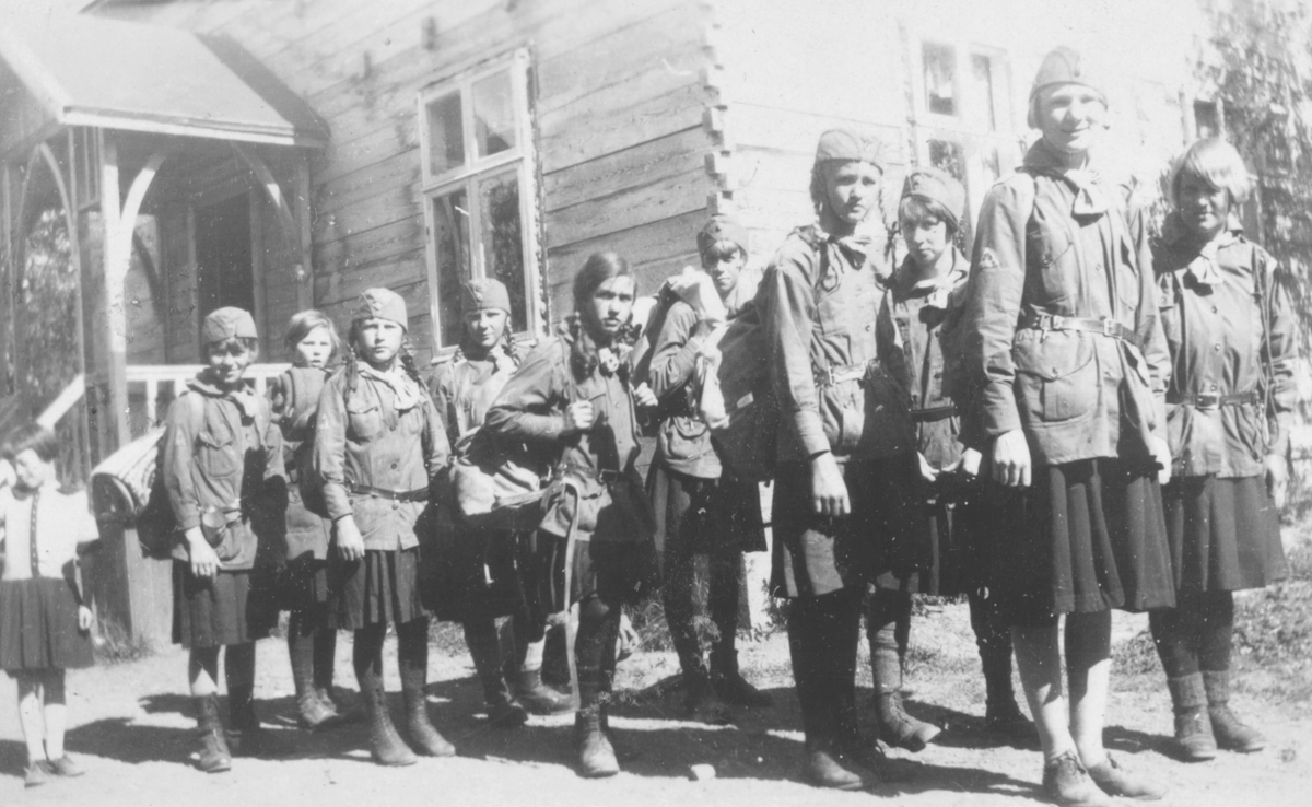 Speiderjenter på patruljeleir i Rustefjelbma fra 4.- 10. juli, 1930. Bildet er fra Kløverpatruljen som står her 'ferdig til start', antakelig til hjemreisen. Tekst fra bildet. Den lille jenta som står til ventre er Anna Andersen. Navn på speiderene er fra venstre: Johanna Dahl, Ragnhild Amble, Randi Feldt, Astrid Pedersen, Arnhild Tøgersen, Aud Johannessen, Ingbjørg Fosslund (Tana), Brit Hornburg, Inger Liv Myreng (Tana), og Vera Hoel. Gjertrud Evanger har tatt bildet. Jentene er kledt i speideruniformer. De bærer på ryggsekker, og utstyr. I bakgrunnen ses huset til Marit Andersen