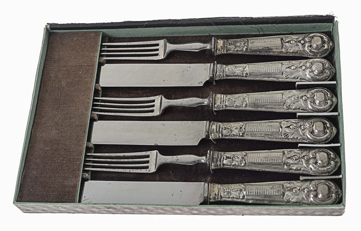 Tre kniver og tre gafler ligger parvis i en eske. Holdt på plass med en tynn bomullstråd. Esken er av delvis stoffbelagt papp. Aldri brukt.