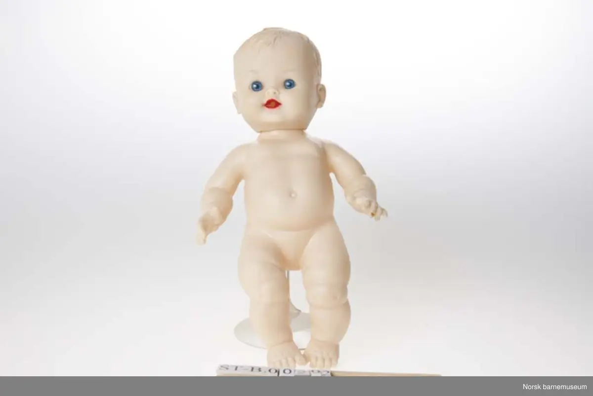 Gutte-dukke i helstøpt figur og hode. 
Har pip i ryggen. Virker ikke. HUdfarget, blå øyne. På malte øyenbryn. Rød munn med hull. Spriker med fingre og storetå. Pipen er rosa og det er "smilehull" i ryggen.